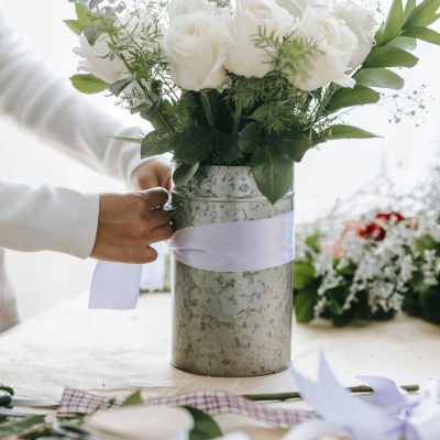 Crop anonymous female florist tying bow on bunch of fresh white roses with decorative branches in floral shop