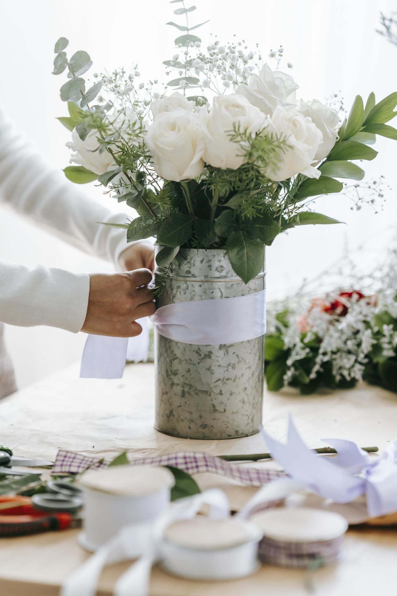 Crop anonymous female florist tying bow on bunch of fresh white roses with decorative branches in floral shop